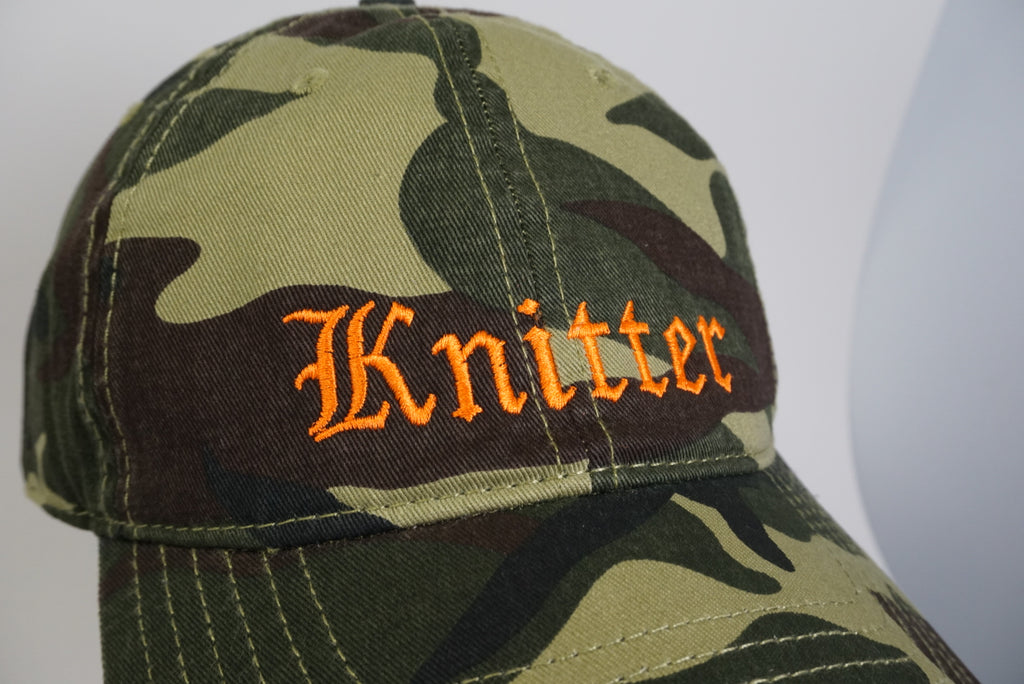Camouflage cap with 'Knitter' embroidered in orange on a light gray background