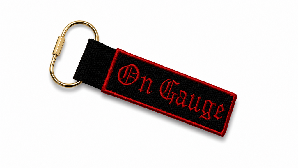 Keychain with 'On Gauge' text on a black strap with a gold clip against a white background