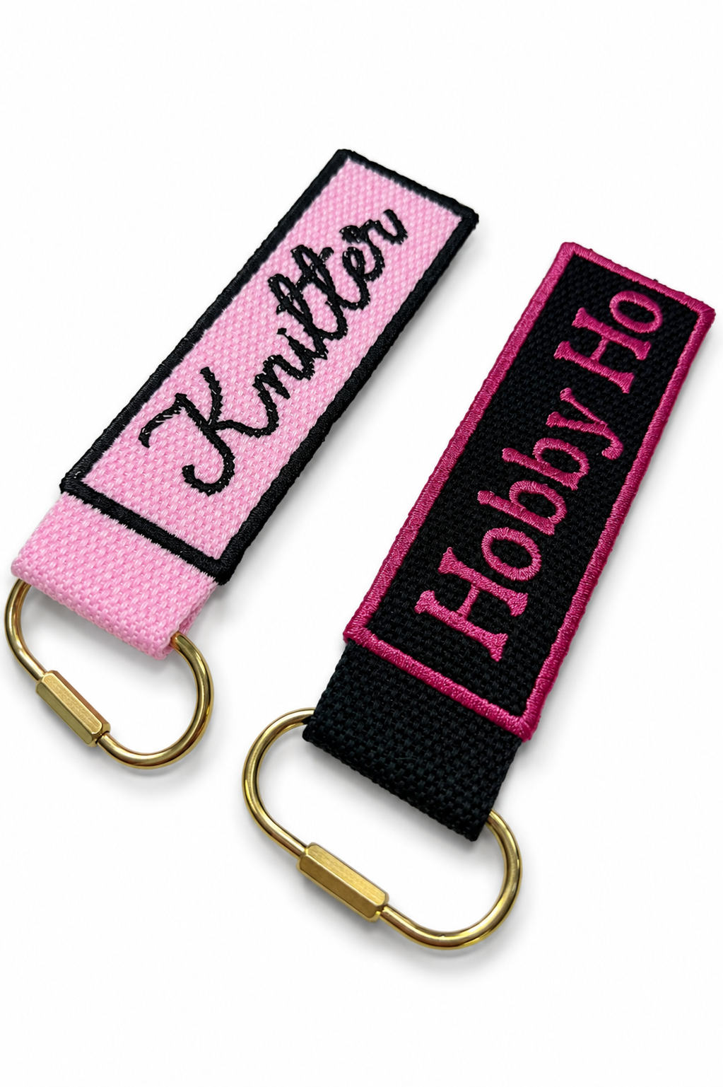 Two keychains with 'Knitter' and 'Hobby Ho' text on pink and black straps with gold carabiners.