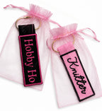 Two keychains with 'Hobby Ho' and 'Knitter' text on pink and black backgrounds, respectively, in gift bags.