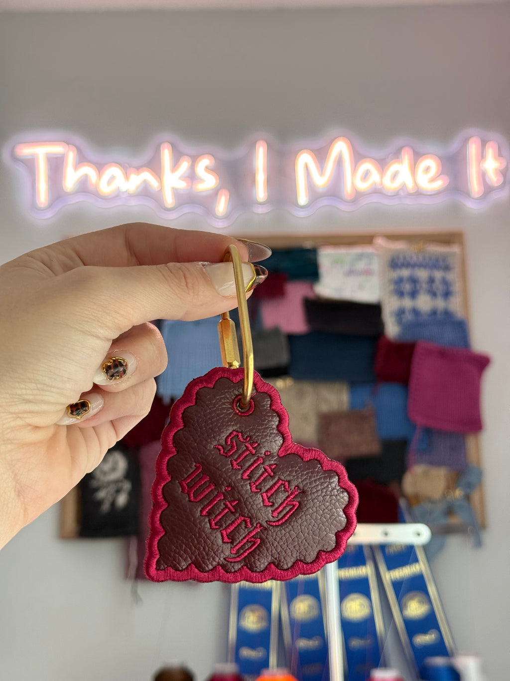 A heart-shaped leather keychain charm with a gold metal clasp. The charm is dark brown textured leather with bright pink embroidered text reading 'Stitch witch' and a matching pink embroidered border. alt