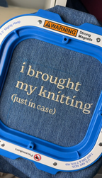 Blue embroidery hoop with text 'i brought my knitting (just in case)' on a denim background.