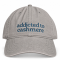 A light gray cotton baseball cap with blue embroidered text reading 'addicted to cashmere' on the front, shown against a plain white background.