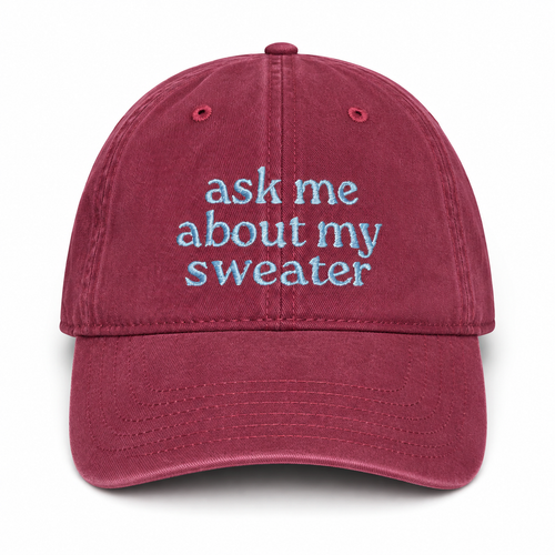 A washed burgundy baseball cap with light blue embroidered text reading 'ask me about my sweater' on the front, shown against a plain white background.