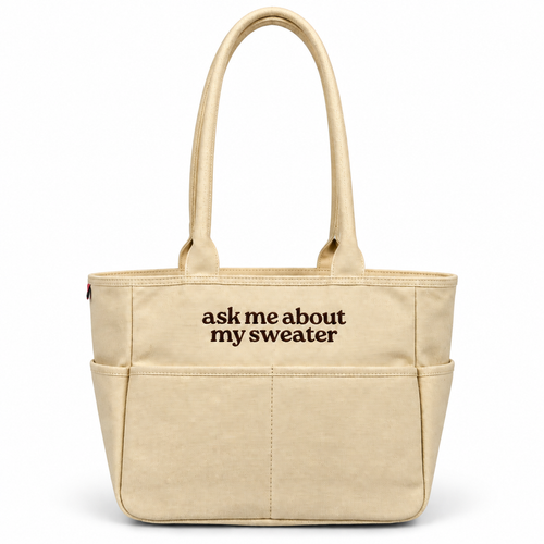 A beige canvas tote bag with two top handles and front pockets, featuring the black printed text 'ask me about my sweater' on the front, set against a plain white background.