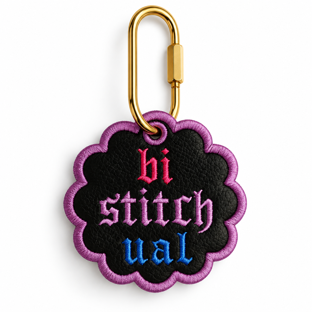 A scalloped black embroidered patch with a purple border and the word 'bi stitchual' in pink, purple, and blue gothic lettering, attached to a gold carabiner on a white background.