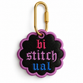 A scalloped black embroidered patch with a purple border and the word 'bi stitchual' in pink, purple, and blue gothic lettering, attached to a gold carabiner on a white background.