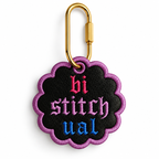 A scalloped black embroidered patch with a purple border and the word 'bi stitchual' in pink, purple, and blue gothic lettering, attached to a gold carabiner on a white background.