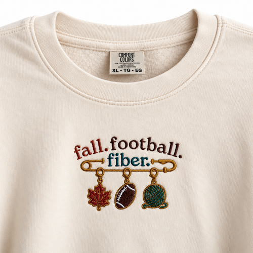 Ivory Comfort Colors sweatshirt with embroidered Fall Football Fiber stitch marker design featuring a leaf, football, and yarn ball.