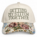 An off-white baseball cap with 'GETTING MY STITCH TOGETHER' embroidered on the front in dark thread, and a floral tapestry pattern on the brim.