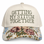 An off-white baseball cap with 'GETTING MY STITCH TOGETHER' embroidered on the front in dark thread, and a floral tapestry pattern on the brim.