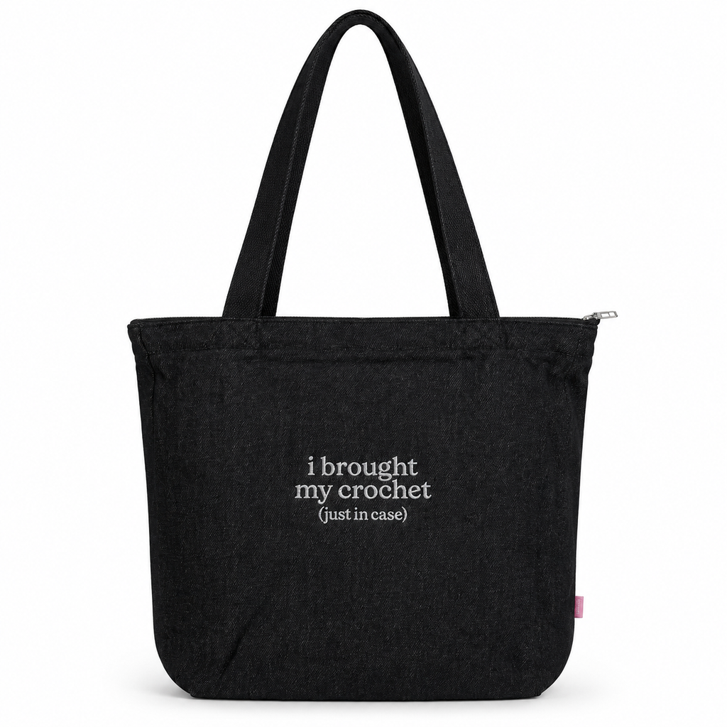 A black canvas tote bag with two shoulder straps, featuring the white printed text 'i brought my crochet (just in case)' on the front, set against a plain white background.