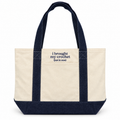 An off-white canvas tote bag with dark navy blue handles, bottom trim, and a front pocket featuring the embroidered text 'i brought my crochet (just in case)'.