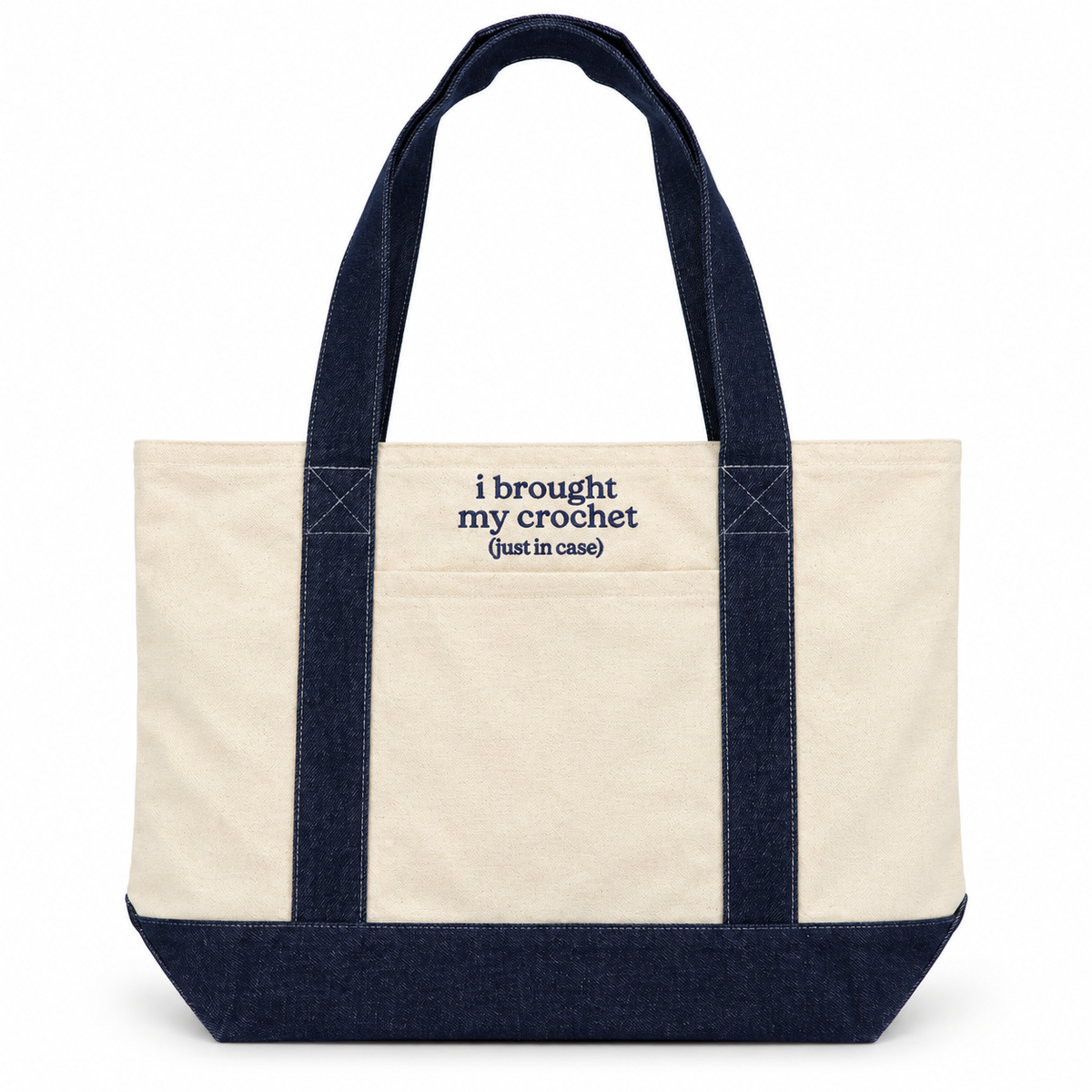 An off-white canvas tote bag with dark navy blue handles, bottom trim, and a front pocket featuring the embroidered text 'i brought my crochet (just in case)'.