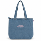 A blue denim tote bag with two shoulder straps, featuring the pink text 'i brought my crochet (just in case)' printed on the front, set against a plain white background.