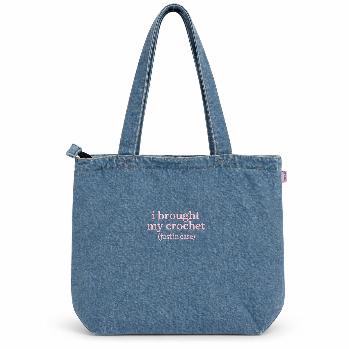 A blue denim tote bag with two shoulder straps, featuring the pink text 'i brought my crochet (just in case)' printed on the front, set against a plain white background.