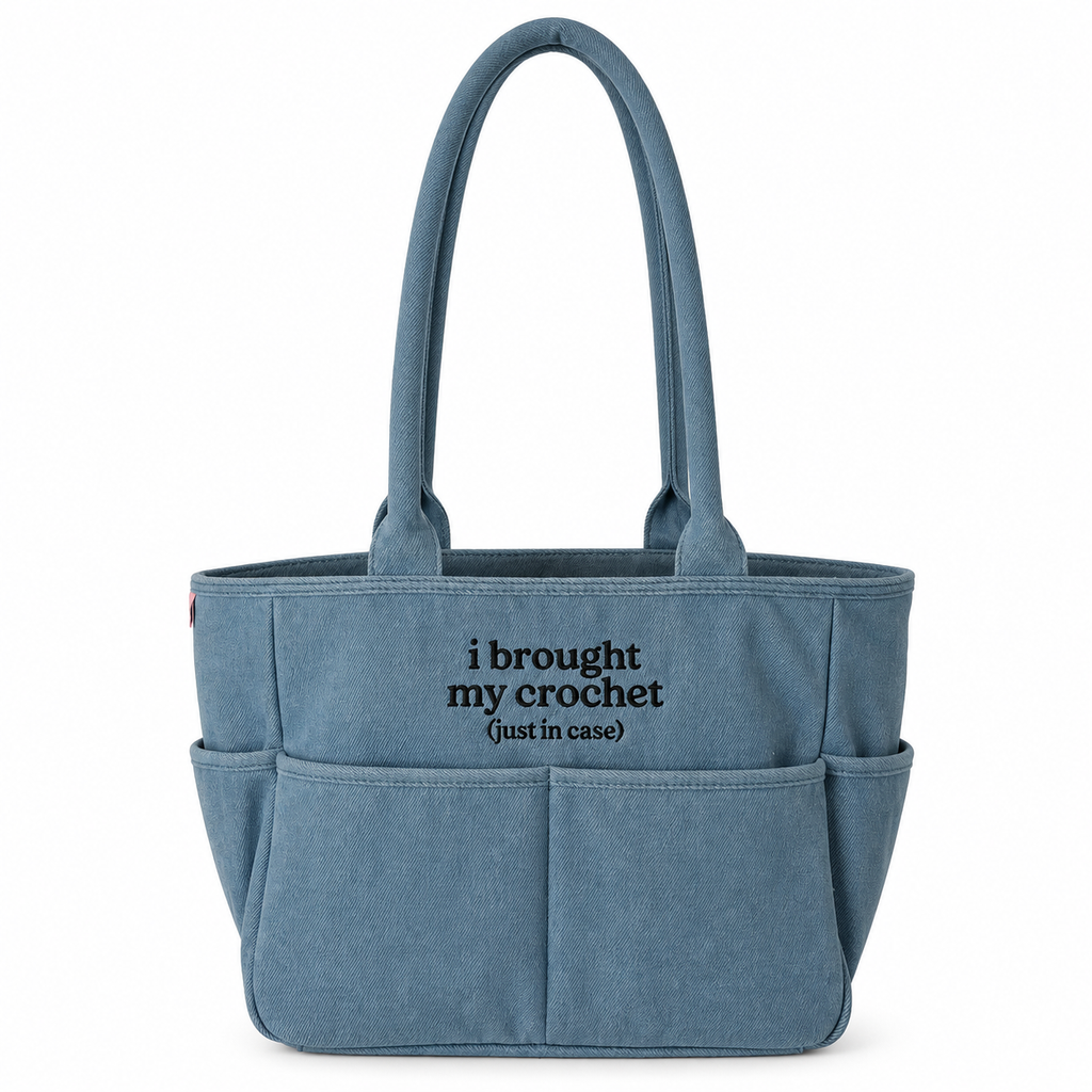 A dusty blue canvas tote bag with dual top handles and front utility pockets, featuring the black printed text 'i brought my crochet (just in case)' on the front, set against a plain white background.