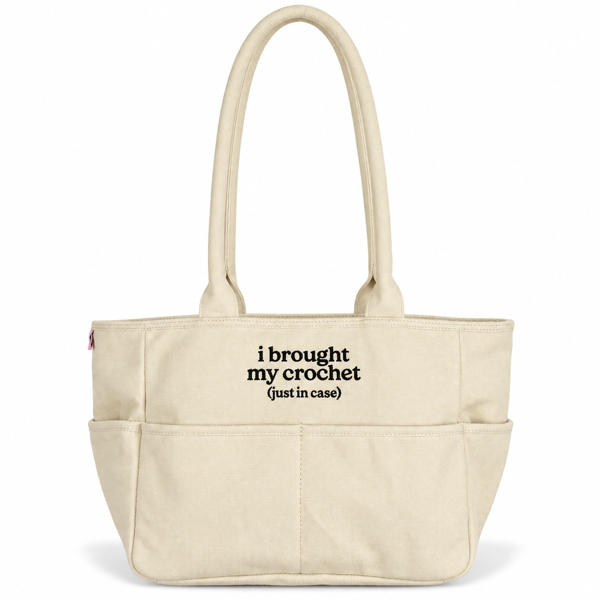 A cream-colored canvas tote bag with black printed text reading 'i brought my crochet (just in case)' on the front pocket, set against a plain white background.