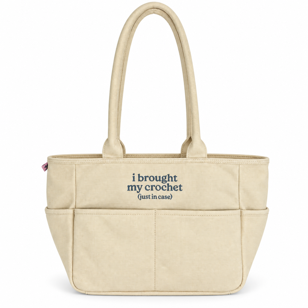 A beige canvas tote bag with two top handles and front pockets, featuring the printed text 'i brought my crochet (just in case)' in navy blue on the front, set against a plain white background.