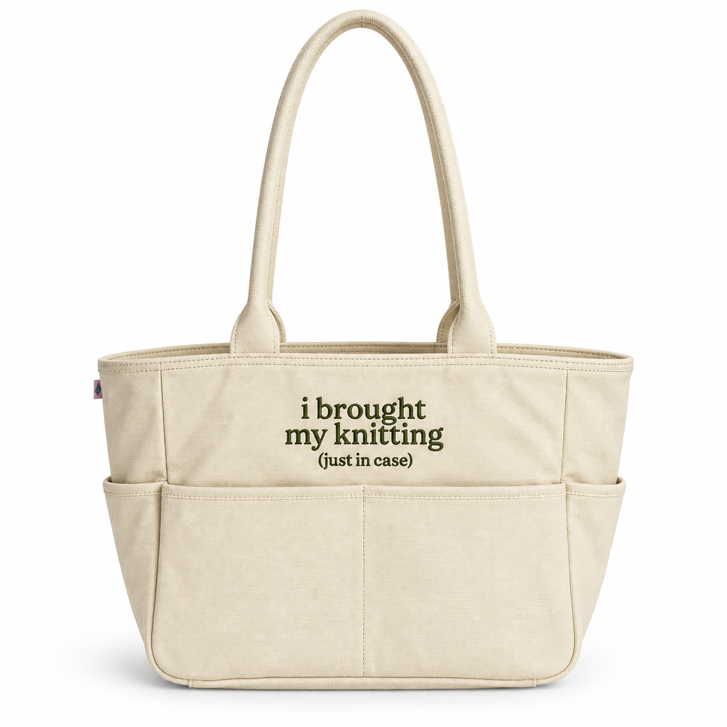 A cream-colored canvas tote bag with two top handles and multiple front pockets. The front of the bag features the printed text 'i brought my knitting (just in case)' in dark olive green.