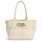 A cream-colored canvas tote bag with two top handles and multiple front pockets. The front of the bag features the printed text 'i brought my knitting (just in case)' in dark olive green.