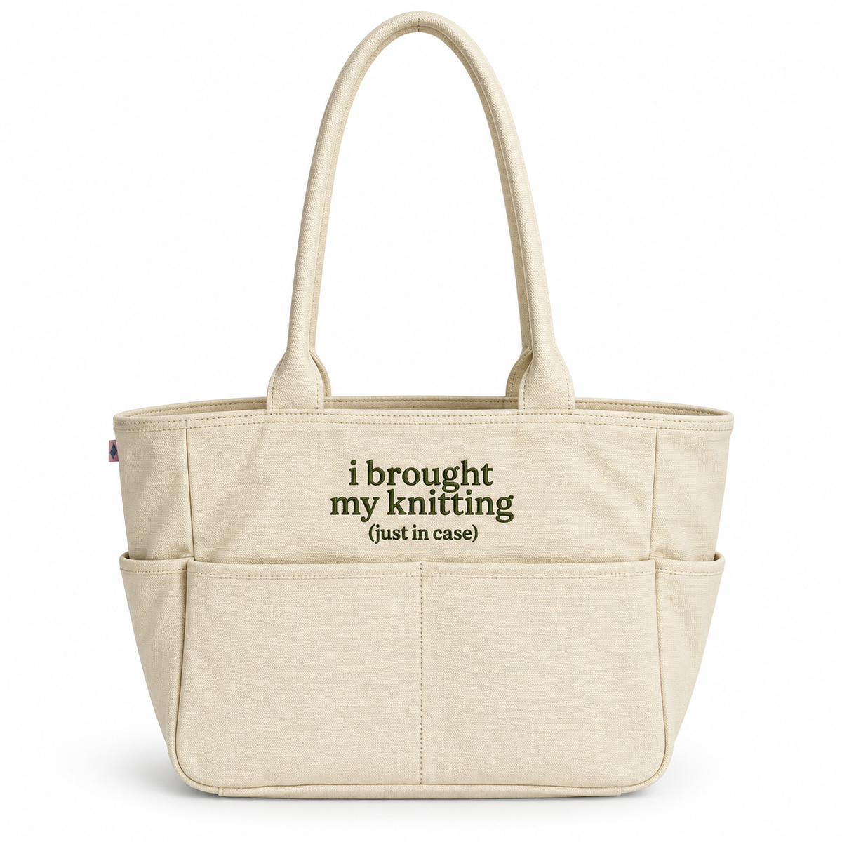 A cream-colored canvas tote bag with two top handles and multiple front pockets. The front of the bag features the printed text 'i brought my knitting (just in case)' in dark olive green.