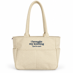 A beige canvas tote bag with two top handles and front pockets, featuring the printed text 'i brought my knitting (just in case)' in navy blue on the front, set against a plain white background.