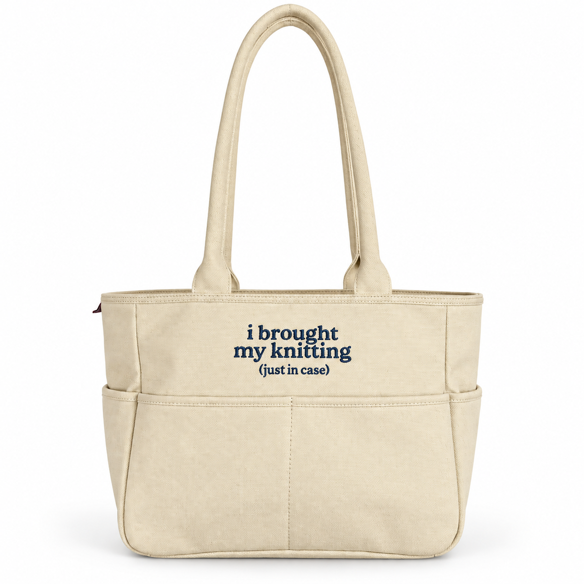 A beige canvas tote bag with two top handles and front pockets, featuring the printed text 'i brought my knitting (just in case)' in navy blue on the front, set against a plain white background.