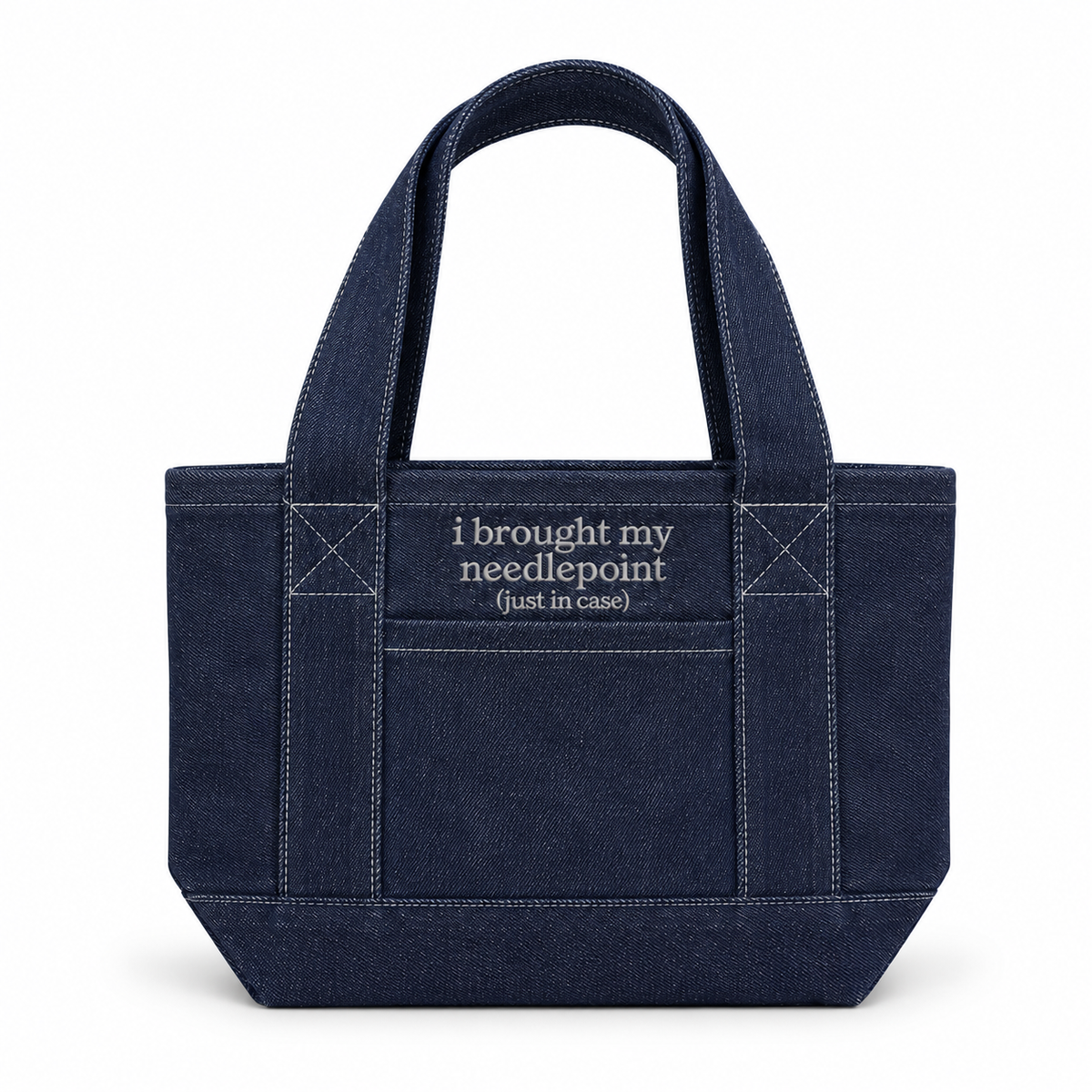 A dark navy blue canvas tote bag with white contrast stitching and white embroidered text on the front pocket that reads 'i brought my needlepoint (just in case)'.