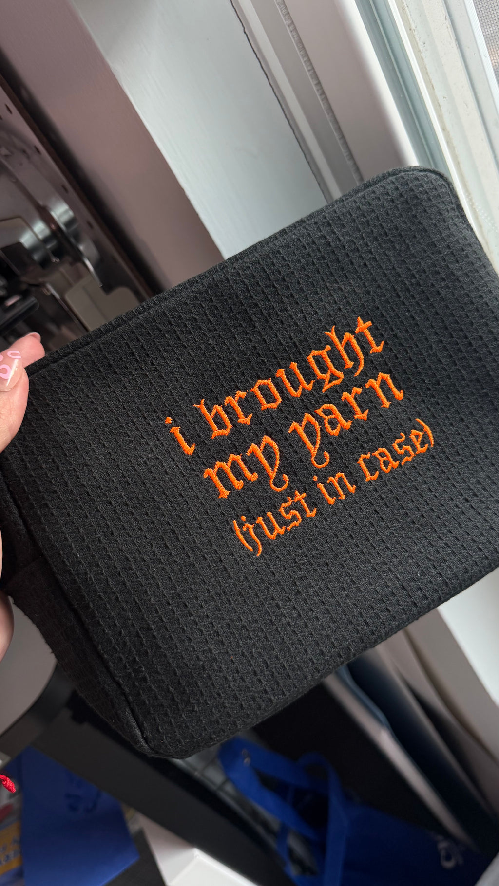 A black textured zippered pouch with orange embroidered text reading 'i brought my yarn (just in case)', held by a hand near a window.