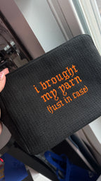 A black textured zippered pouch with orange embroidered text reading 'i brought my yarn (just in case)', held by a hand near a window.