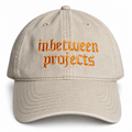 A beige baseball cap with 'inbetween projects' embroidered in orange gothic-style lettering on the front, shown against a plain white background.