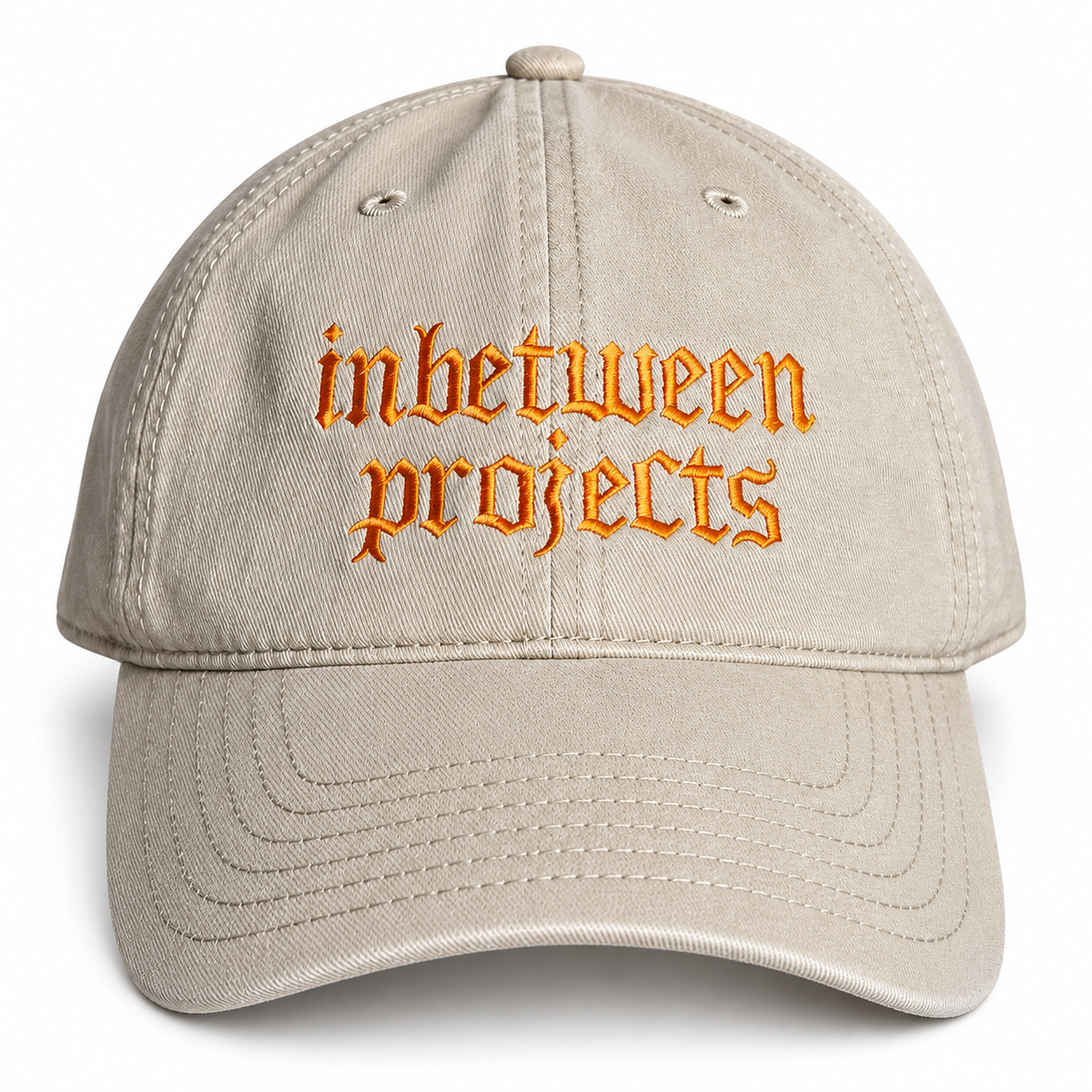 A beige baseball cap with 'inbetween projects' embroidered in orange gothic-style lettering on the front, shown against a plain white background.