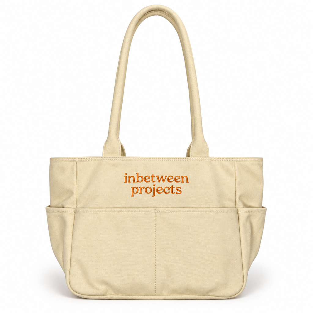 A beige canvas tote bag with two top handles and a front pocket, featuring the words 'inbetween projects' printed in orange on the front, set against a plain white background.