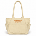 A beige canvas tote bag with two top handles and a front pocket, featuring the words 'inbetween projects' printed in orange on the front, set against a plain white background.