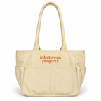 A beige canvas tote bag with two top handles and a front pocket, featuring the words 'inbetween projects' printed in orange on the front, set against a plain white background.