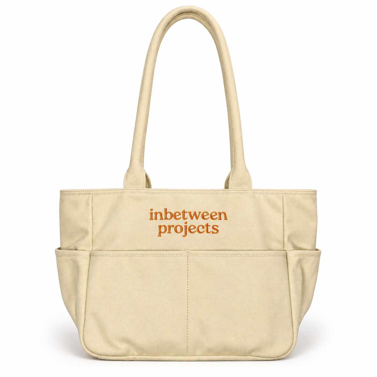 A beige canvas tote bag with two top handles and a front pocket, featuring the words 'inbetween projects' printed in orange on the front, set against a plain white background.