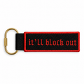Black bag charm with red border and red gothic “It’ll Block Out” embroidery, attached to a gold clip.