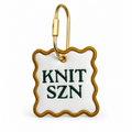 White faux leather bag tag with green Knit SZN embroidery, old gold border, and gold-tone hardware