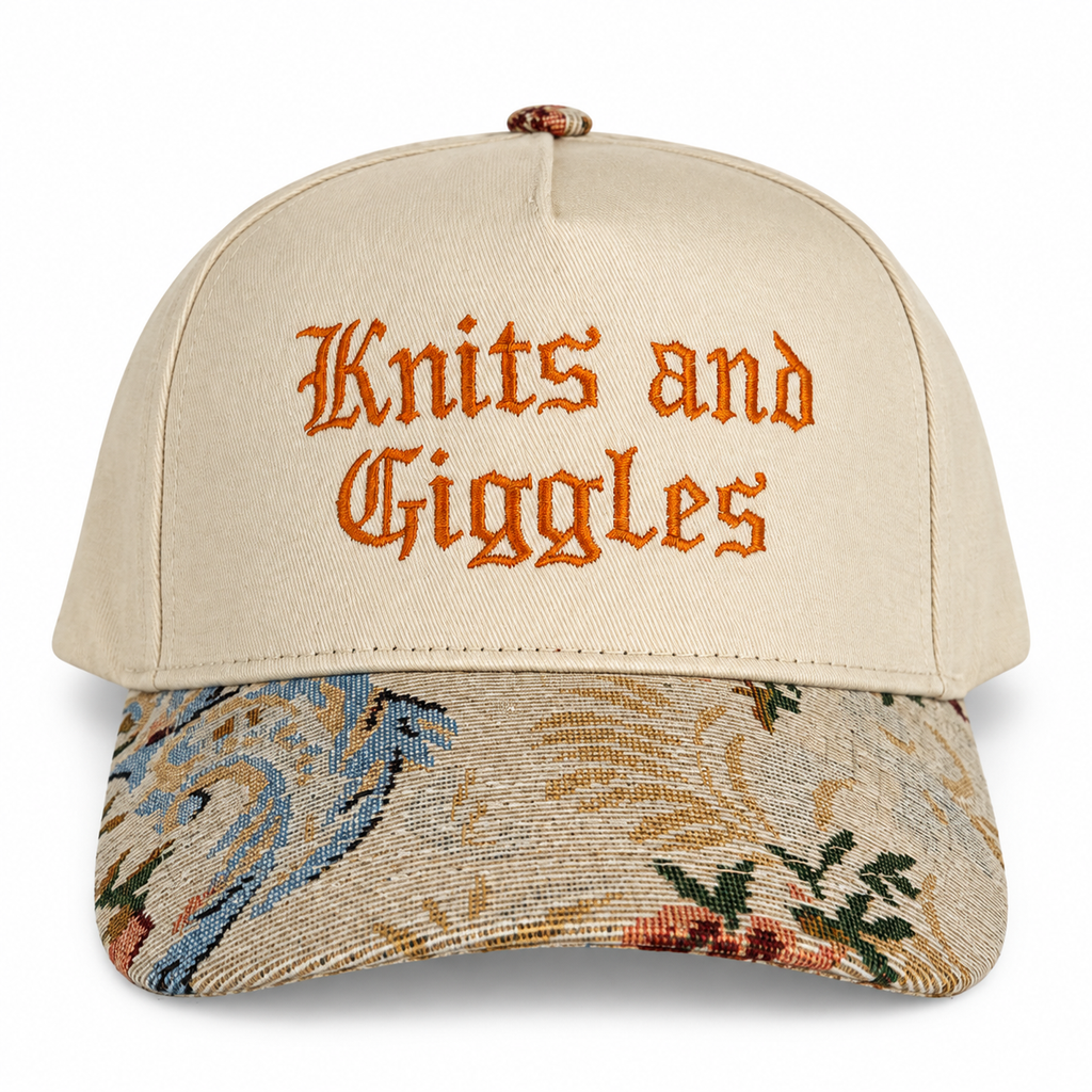 A beige baseball cap with 'Knits and Giggles' embroidered in orange gothic lettering on the front. The curved brim features a detailed, multicolored woven floral pattern in beige, blue, green, and orange.
