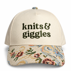 An off-white baseball cap with 'knits & giggles' embroidered in dark olive green on the front. The curved brim features a detailed floral and paisley tapestry pattern in beige, blue, red, and green.