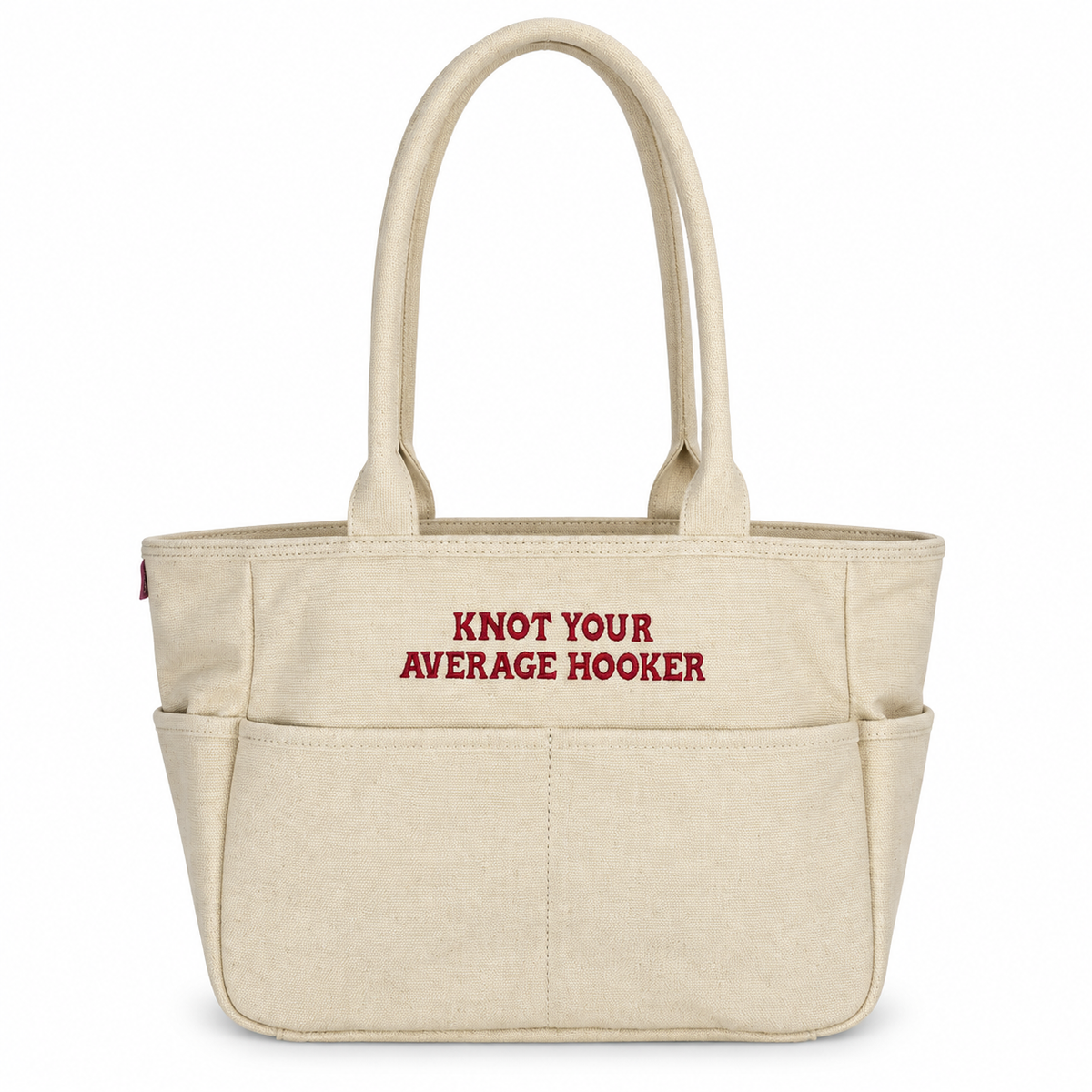 A natural canvas tote bag with two top handles and front pockets, featuring the red embroidered text 'KNOT YOUR AVERAGE HOOKER' on the front, set against a plain white background.