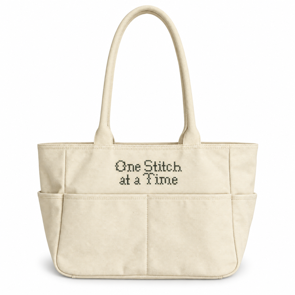 Natural canvas TIMI Tote with dark olive “One Stitch at a Time” cross stitch embroidery.