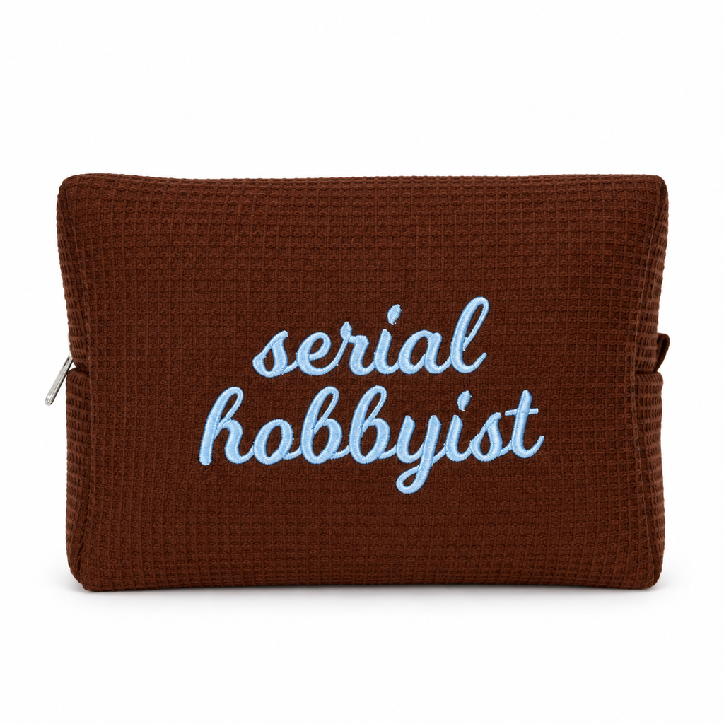 A brown waffle-weave zip pouch with the words 'serial hobbyist' embroidered in light blue script on the front, shown against a plain white background.