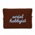 A brown waffle-weave zip pouch with the words 'serial hobbyist' embroidered in light blue script on the front, shown against a plain white background.