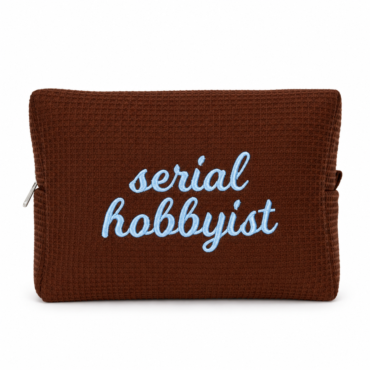 A brown waffle-weave zip pouch with the words 'serial hobbyist' embroidered in light blue script on the front, shown against a plain white background.