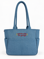 A blue fabric tote bag with dual shoulder straps, front utility pockets, and the text 'spiritually on gauge' embroidered in red and yellow on the front, shown against a plain white background.