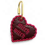 A heart-shaped leather keychain charm with a gold metal clasp. The charm is dark brown textured leather with bright pink embroidered text reading 'Stitch witch' and a matching pink embroidered border.