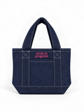Dark denim mini tote with bright pink “Work in Progress” embroidery.