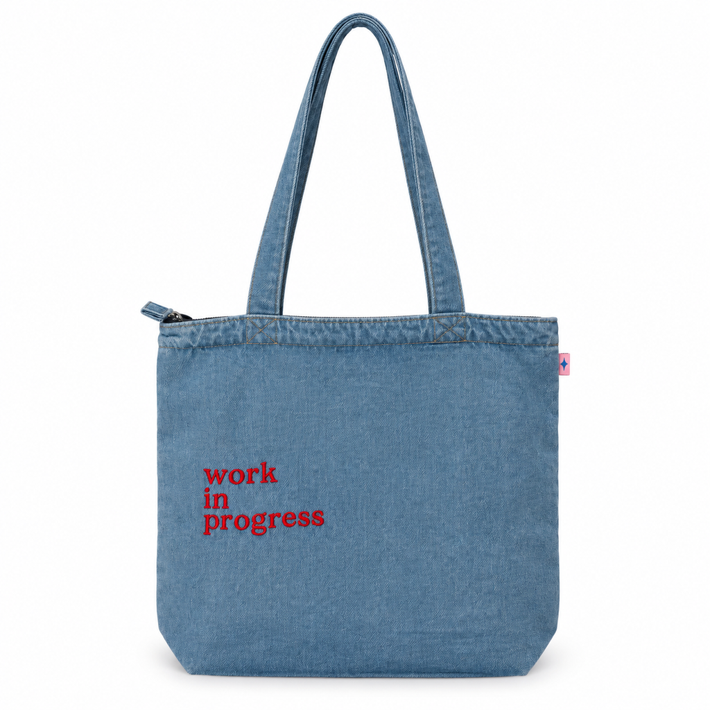 A blue denim tote bag with 'work in progress' printed in red text on the front, shown against a plain white background.